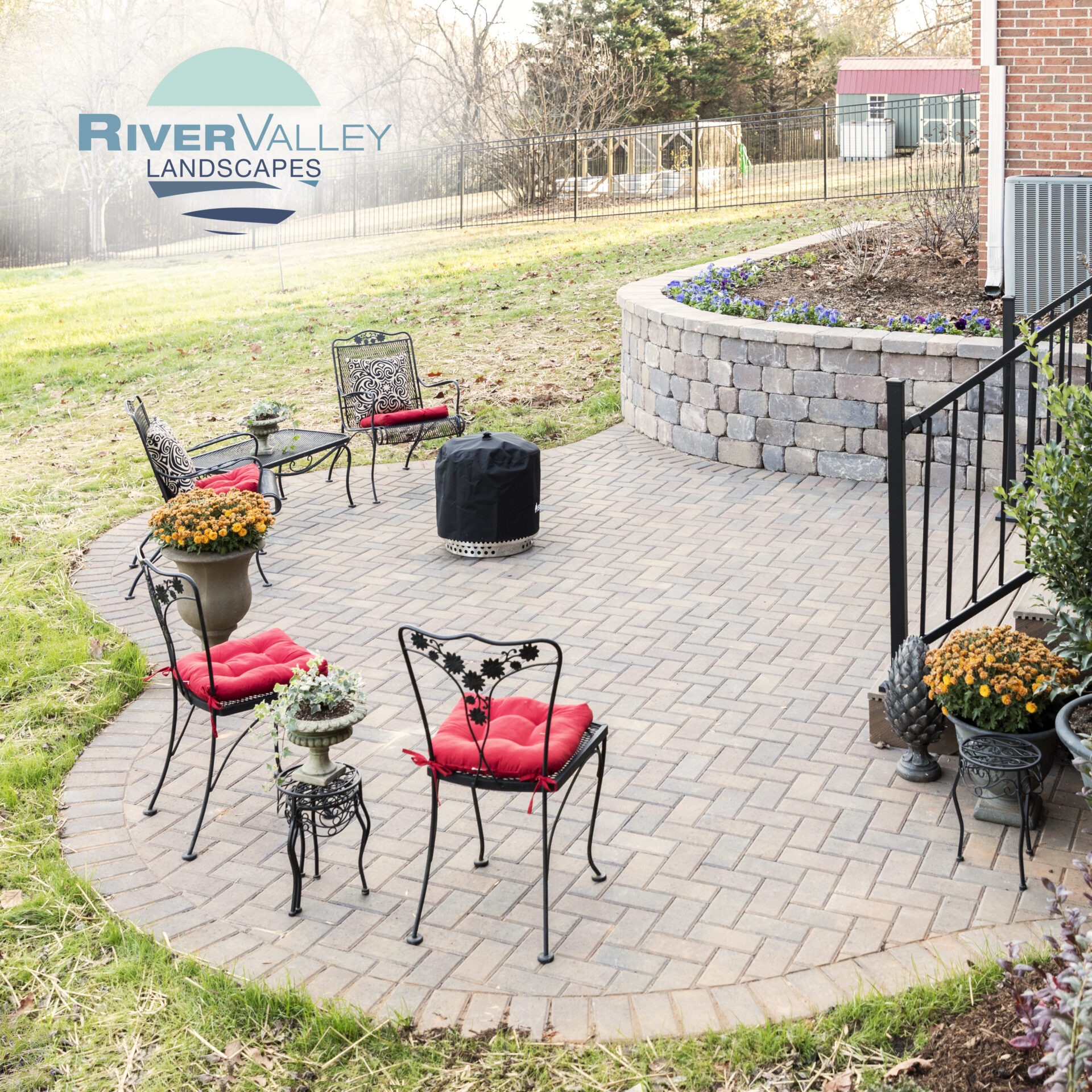A landscaped patio features red-cushioned chairs, potted flowers, and a small garden wall, set in a grassy yard with fenced surroundings.