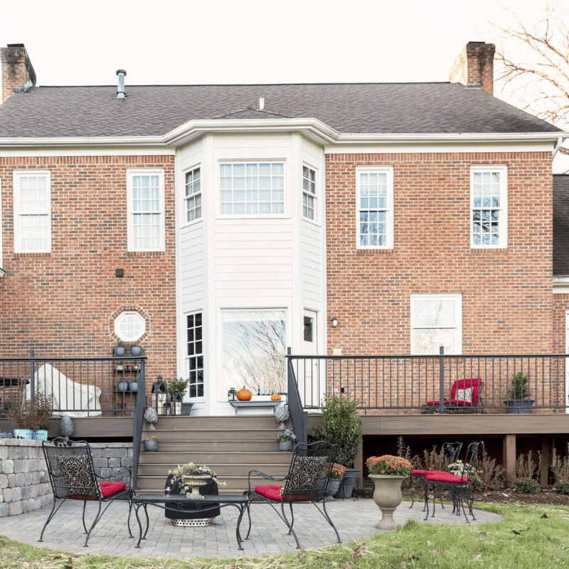 Brick house with wooden deck, patio furniture, and potted plants. No visible people or landmarks. Peaceful outdoor setting with a garden backdrop.