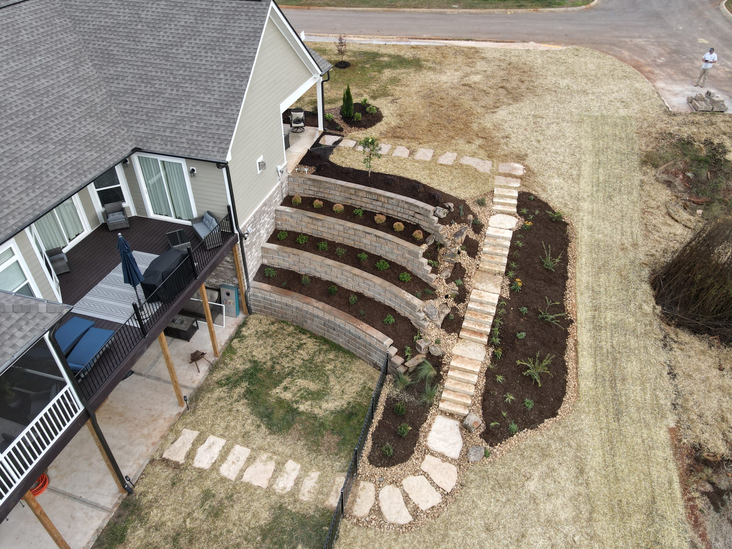 Aerial view of house with tiered garden, stone path, patio furniture, and a person standing near driveway. Grass and mulch surrounding landscape.