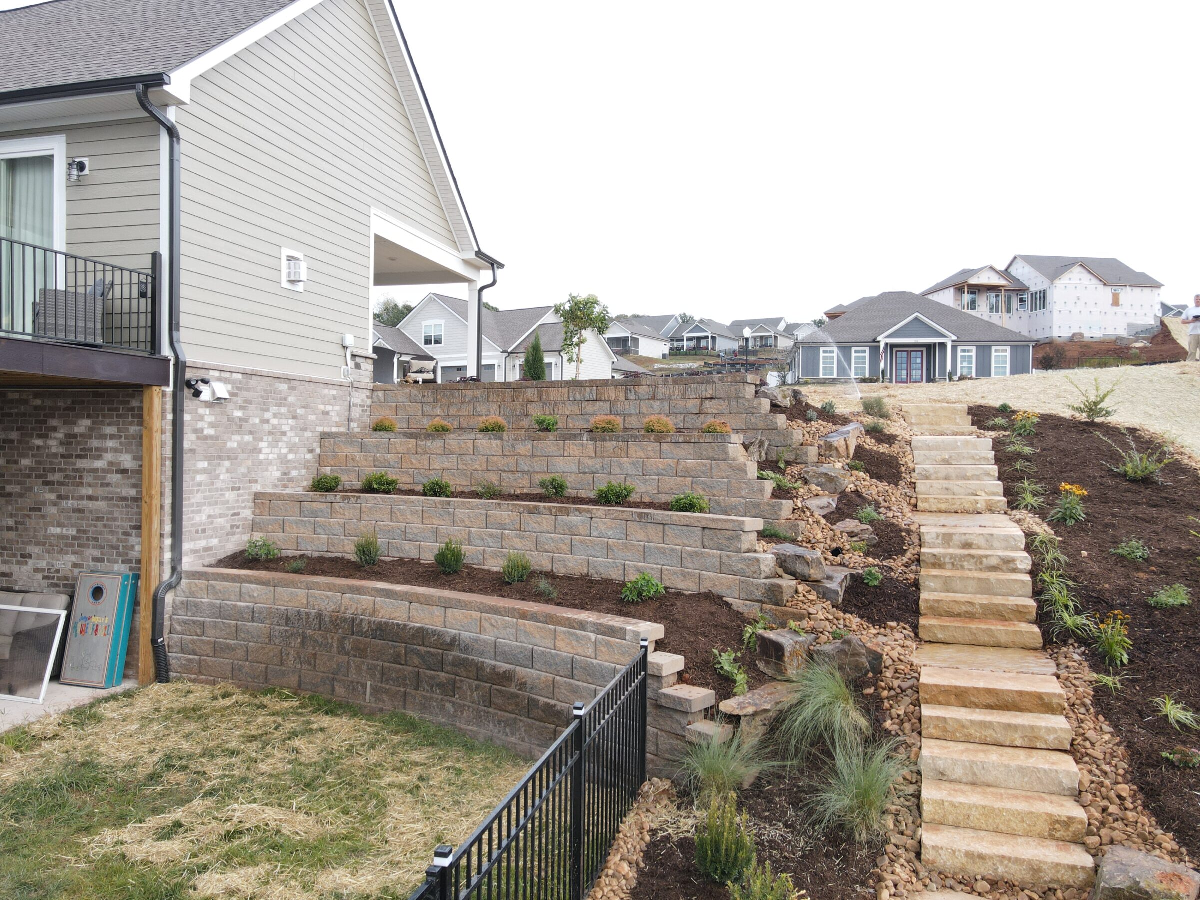 Terraced backyard with stone retaining walls and steps, surrounded by newly planted shrubs. Suburban neighborhood with modern houses visible in the background.