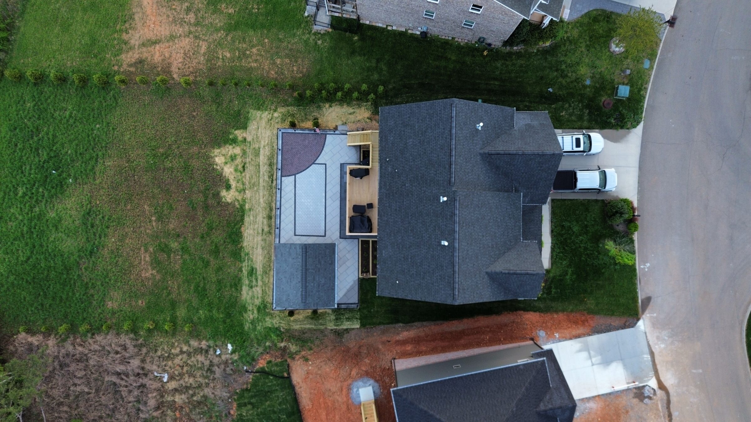 Aerial view of a house with a patio, driveway, and two cars parked. Surrounded by green lawns and nearby roads.