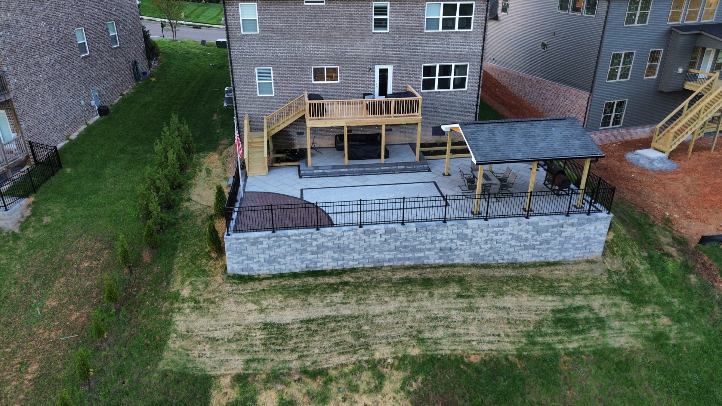 A backyard features a raised patio with a wooden deck and gazebo, surrounded by a stone wall and grassy yard.