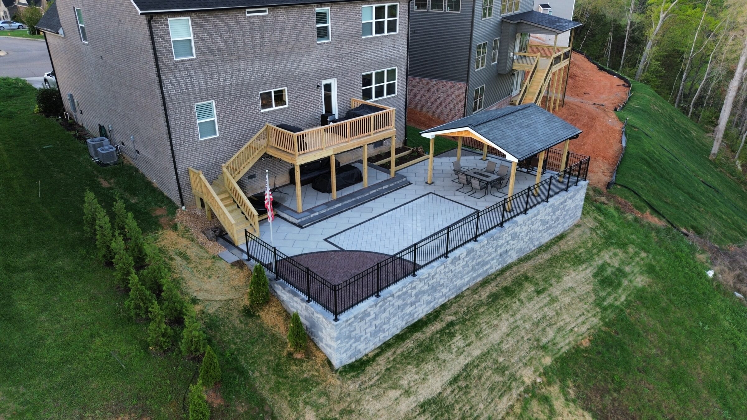 Modern house with a spacious elevated patio, covered seating area, and flagpole, overlooking green slopes and a fenced backyard.