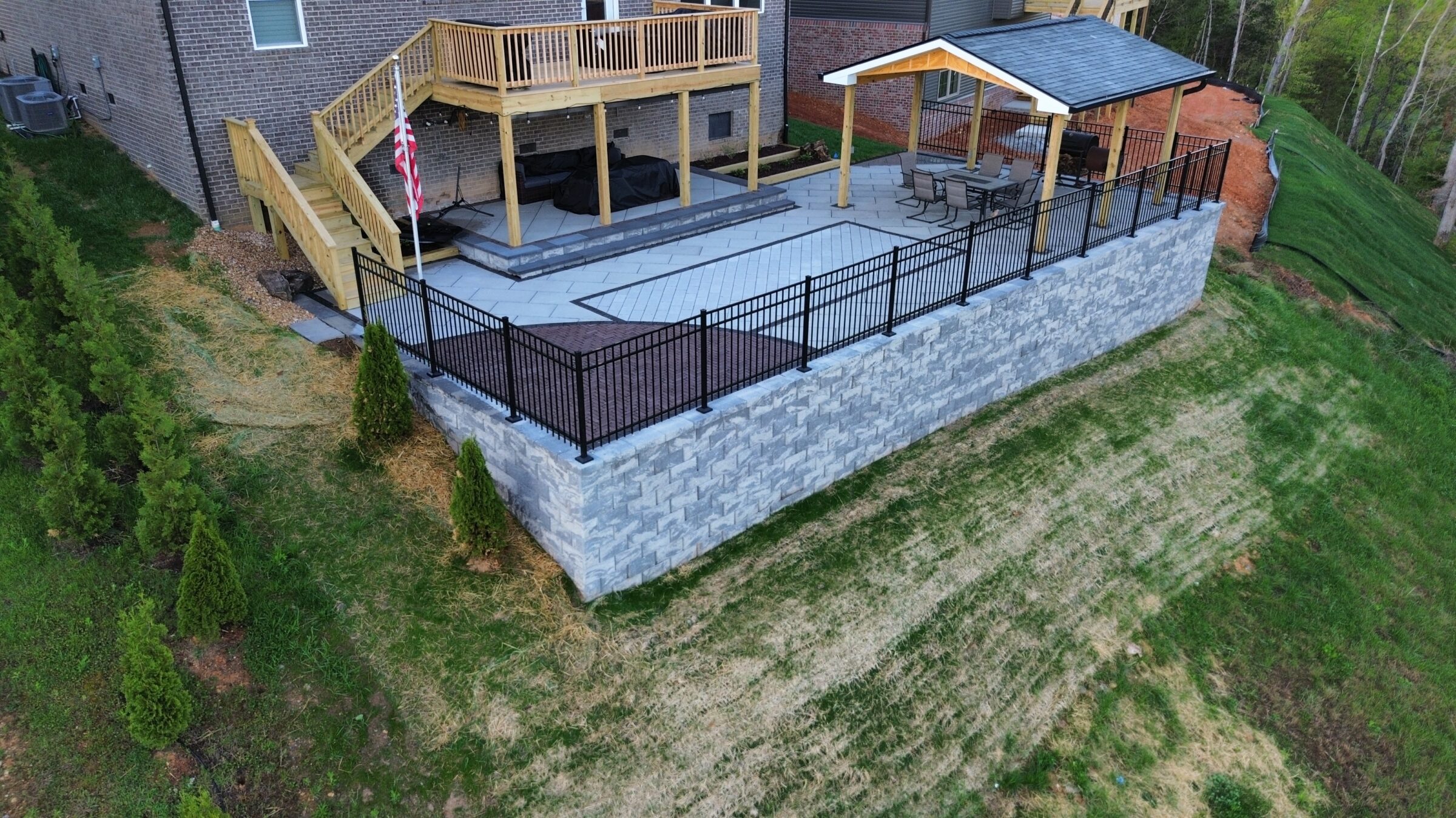 Elevated backyard patio with wooden deck, covered seating area, and flag. Surrounded by a metal fence, brick wall, and lush greenery.