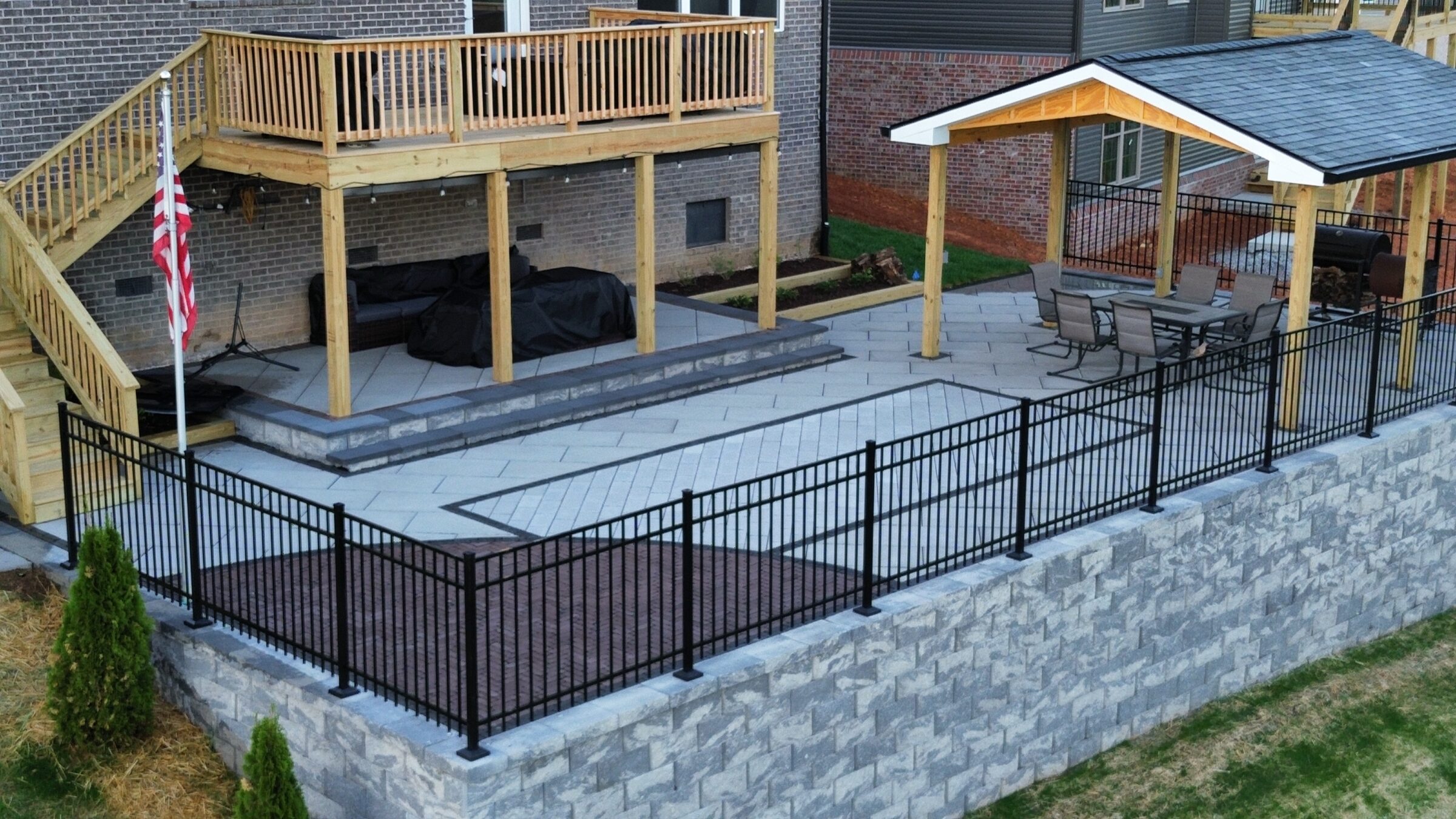 Elevated patio with wooden deck, American flag, and seating area under a pergola. Fenced with stone wall, adjacent to a brick house.