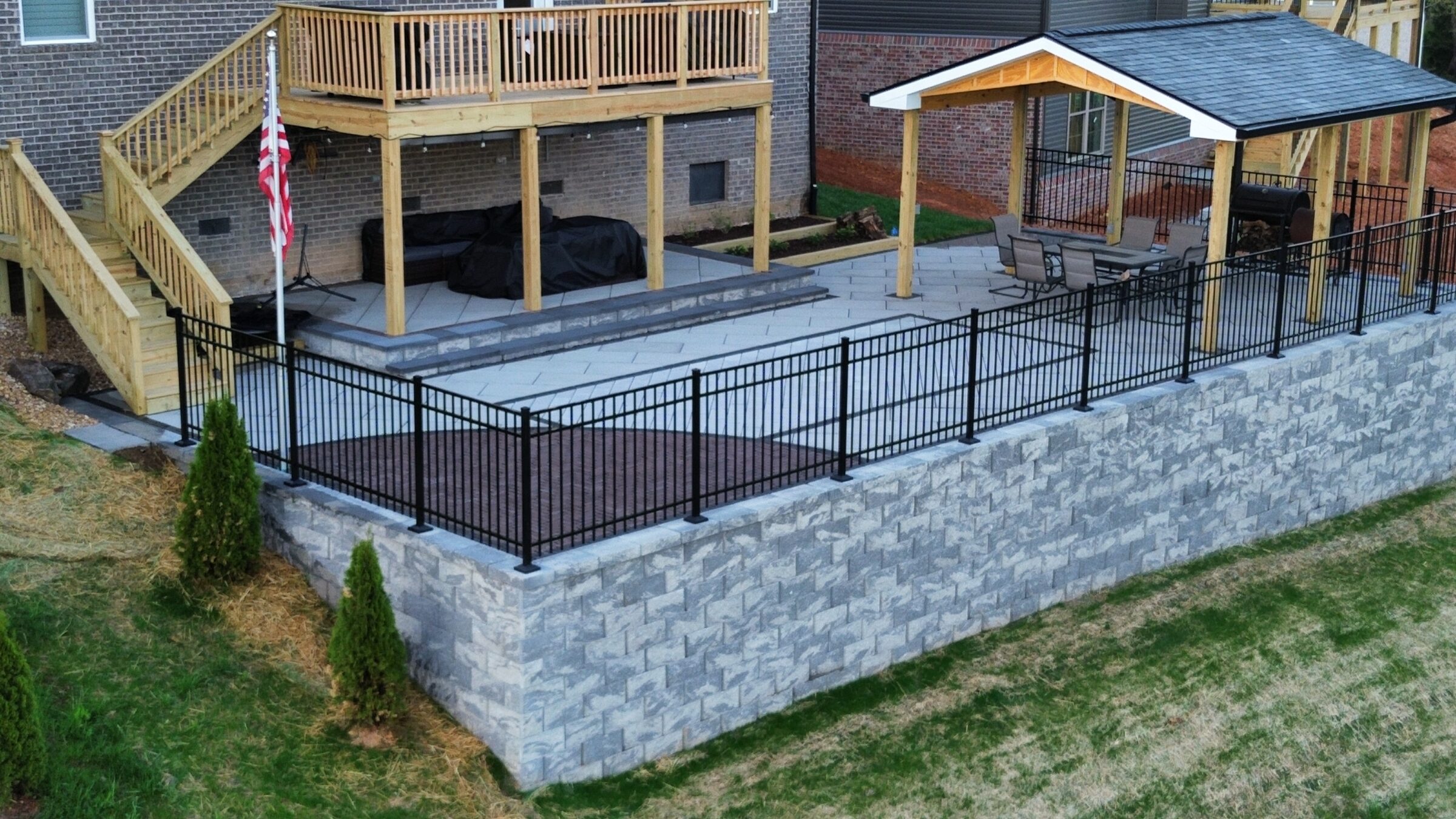 A backyard patio with a wooden deck, stone wall, and flagpole. Covered seating area beneath roof, surrounded by a black metal fence.