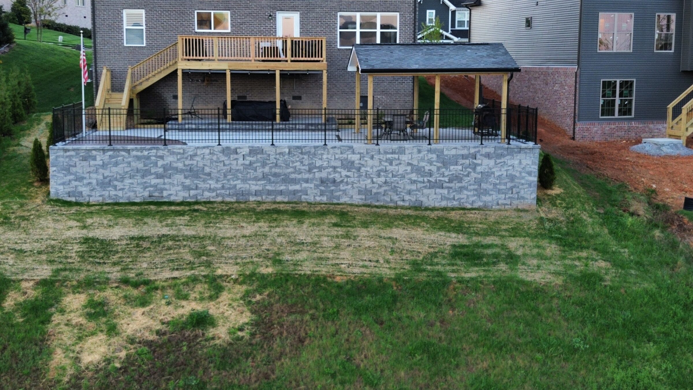 Brick house with wooden deck and covered patio, fenced yard with retaining wall, and visible American flag.