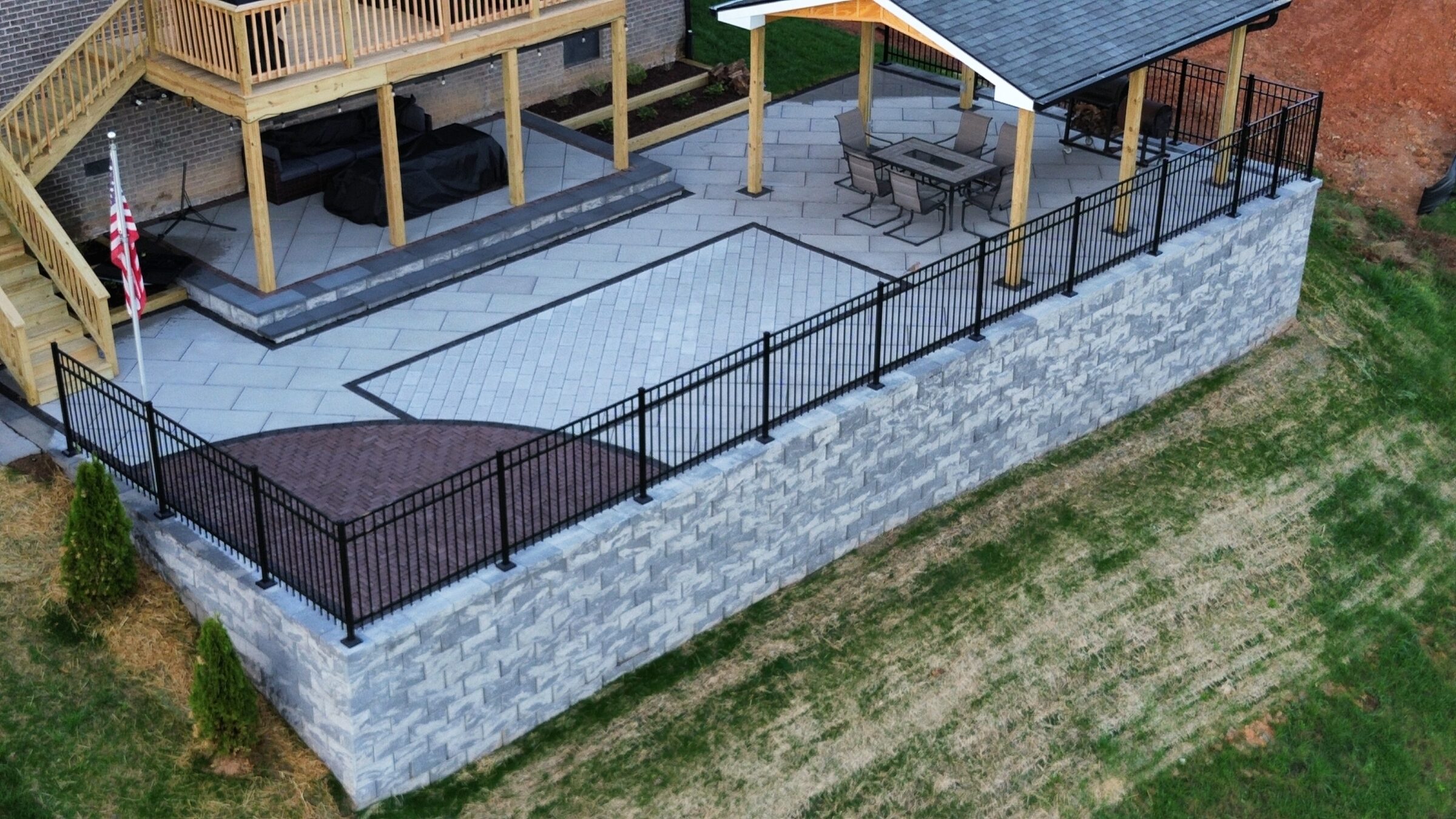 Elevated patio with a roofed area, wooden deck, flagpole, and modern railing, surrounded by landscaped grass and retaining wall.