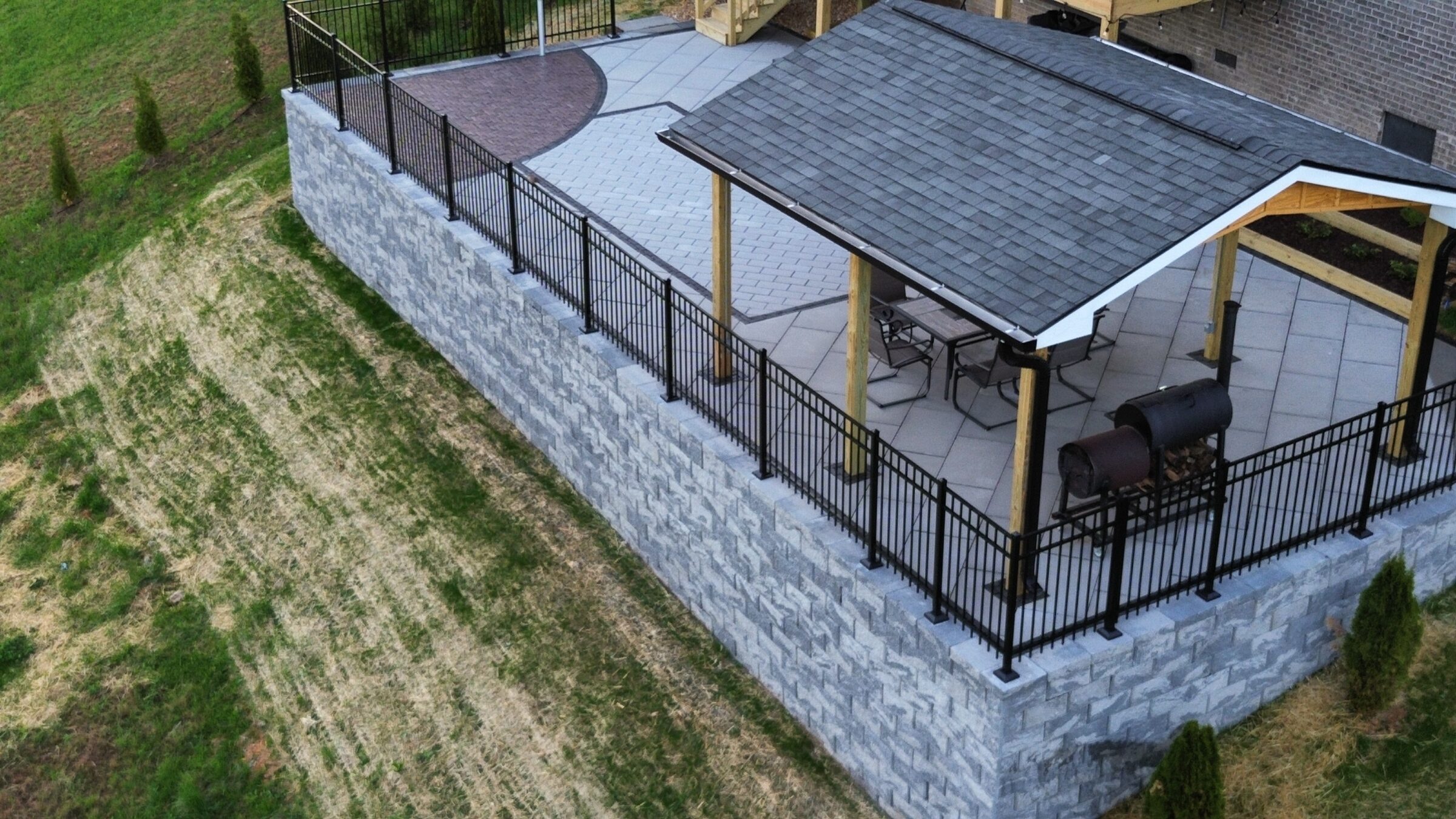 A covered patio sits on a raised area with black railings, featuring outdoor furniture and a grill, surrounded by greenery.