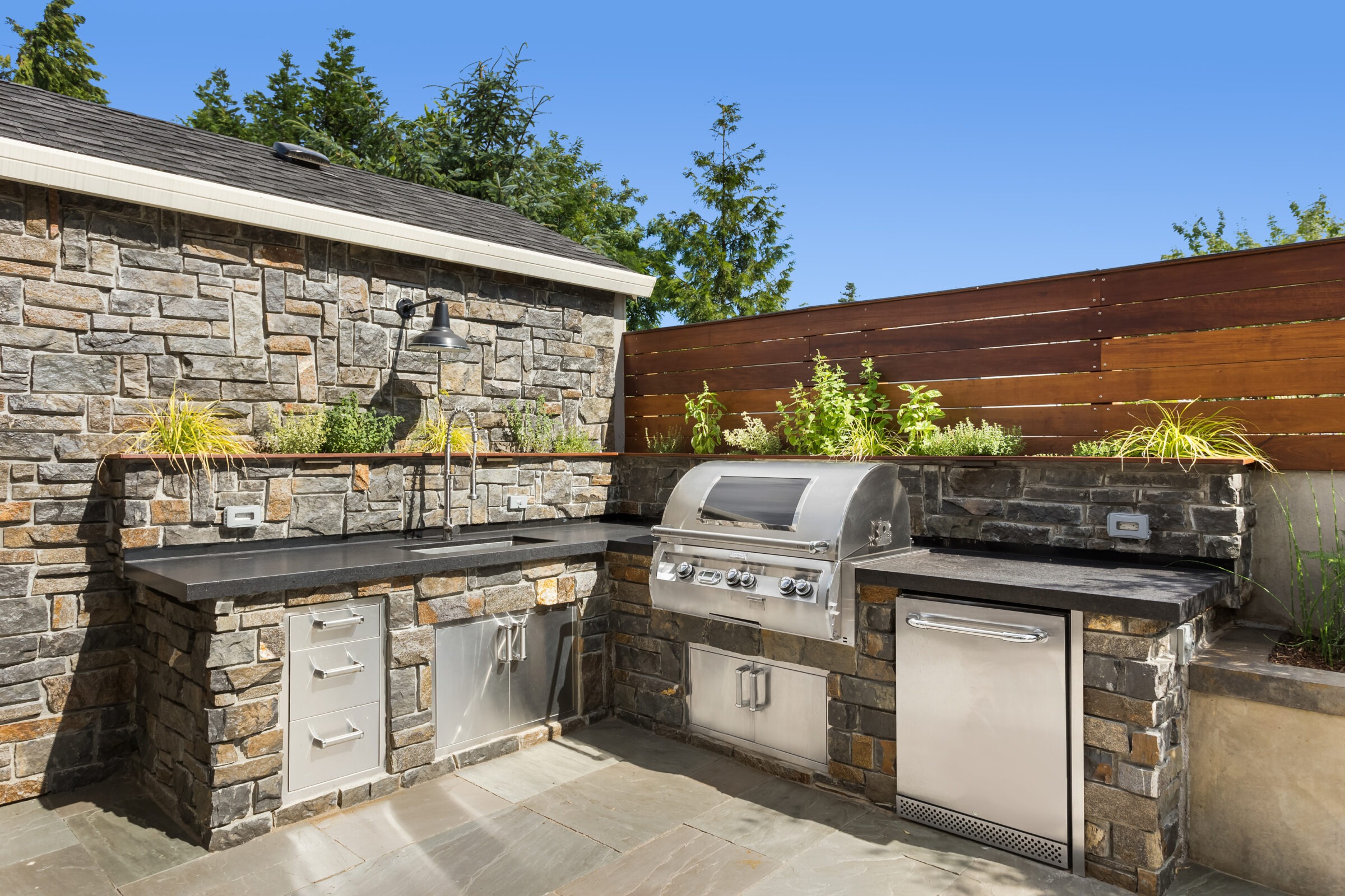 Outdoor kitchen with stone walls features a built-in grill, sink, and stainless steel cabinets, surrounded by plants and wooden fencing.