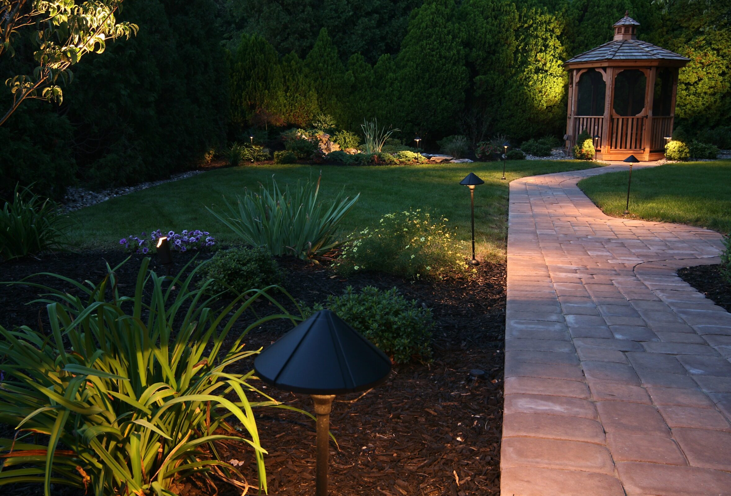 A serene garden path with lush greenery and a wooden gazebo is softly illuminated by landscape lighting at dusk.