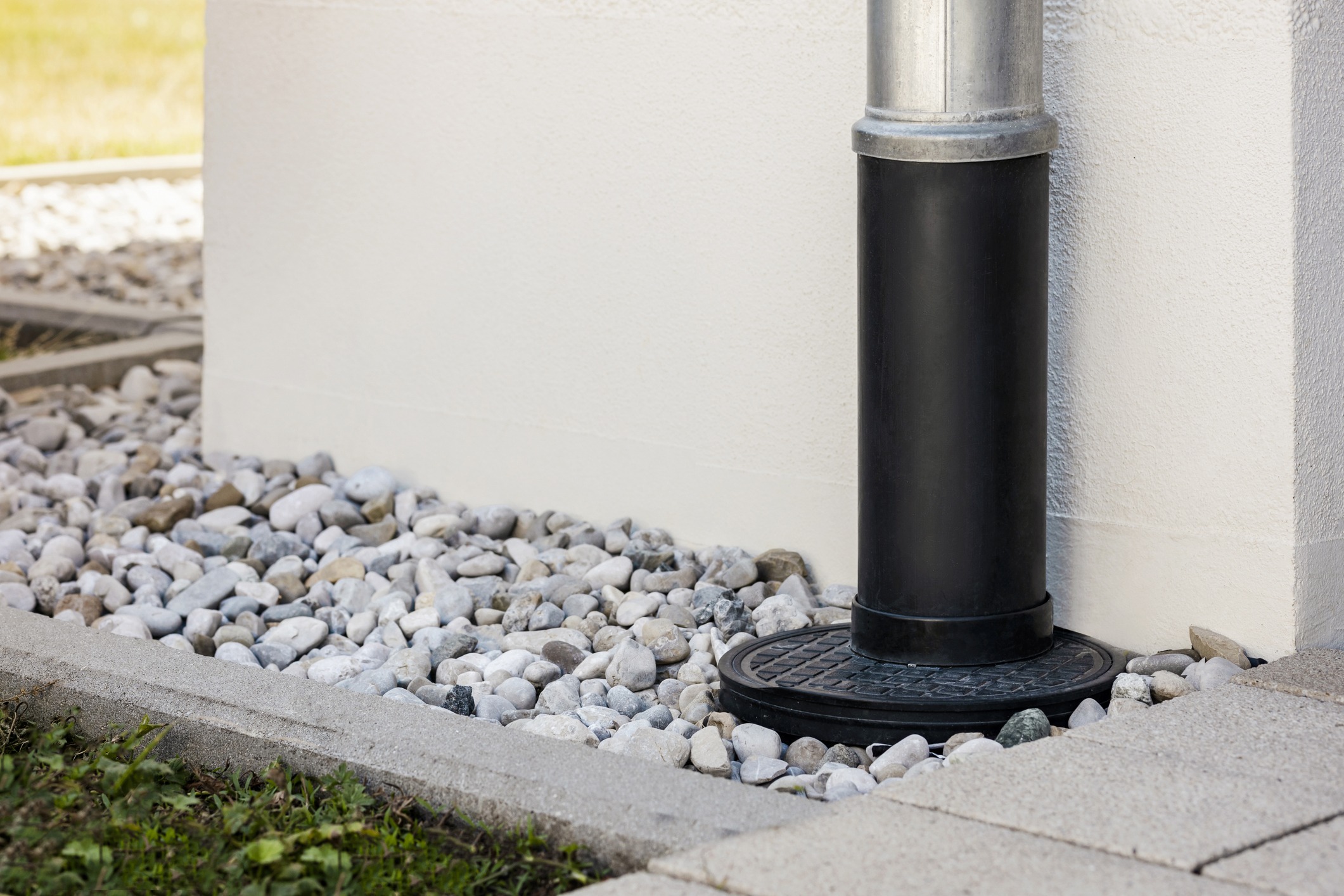 A black drainage pipe against a white wall, surrounded by light-colored pebbles and concrete edging, with green grass nearby.
