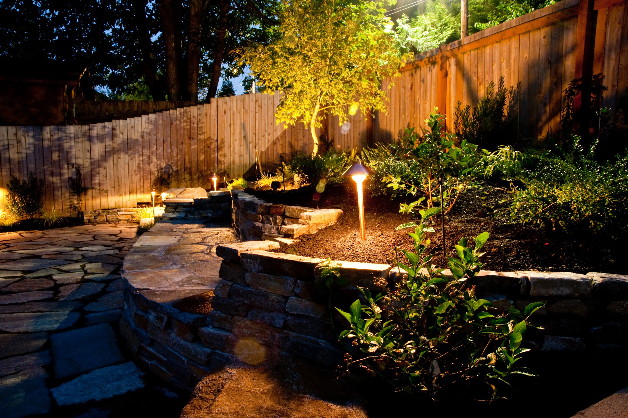 A landscaped garden at dusk features stone pathways, wooden fences, manicured plants, and warm pathway lights illuminating the serene outdoor setting.